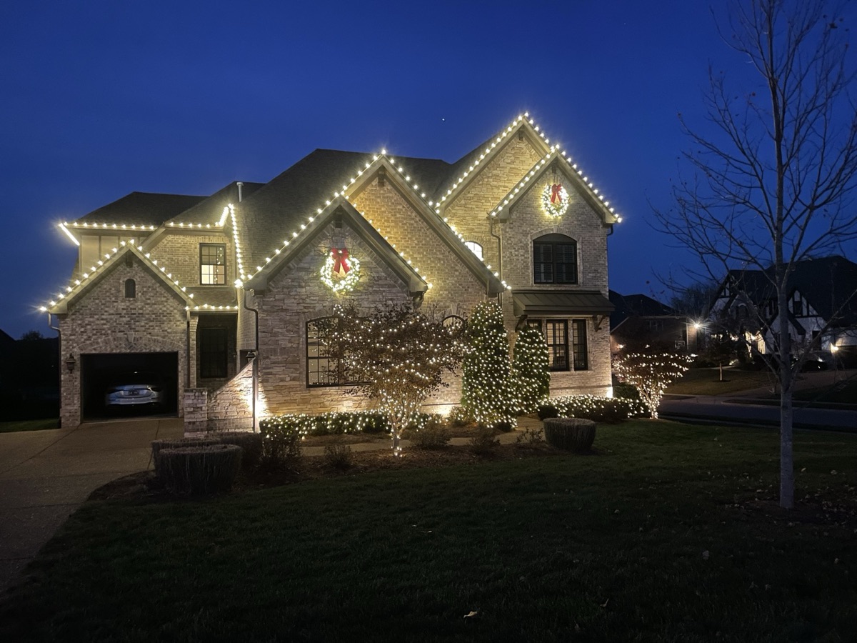 Two-story brick home with warm white rooflines, lit wreaths with red bows, and wrapped trees