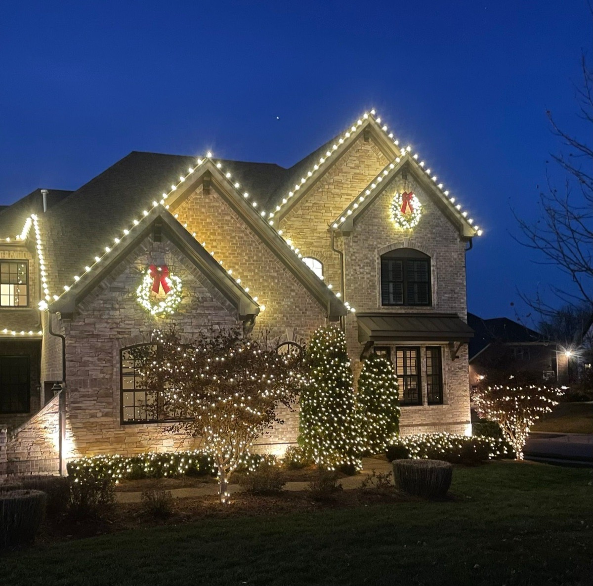 Brick home at blue hour with warm white rooflines, lit wreaths with red bows on both peaks, and wrapped trees and shrubs